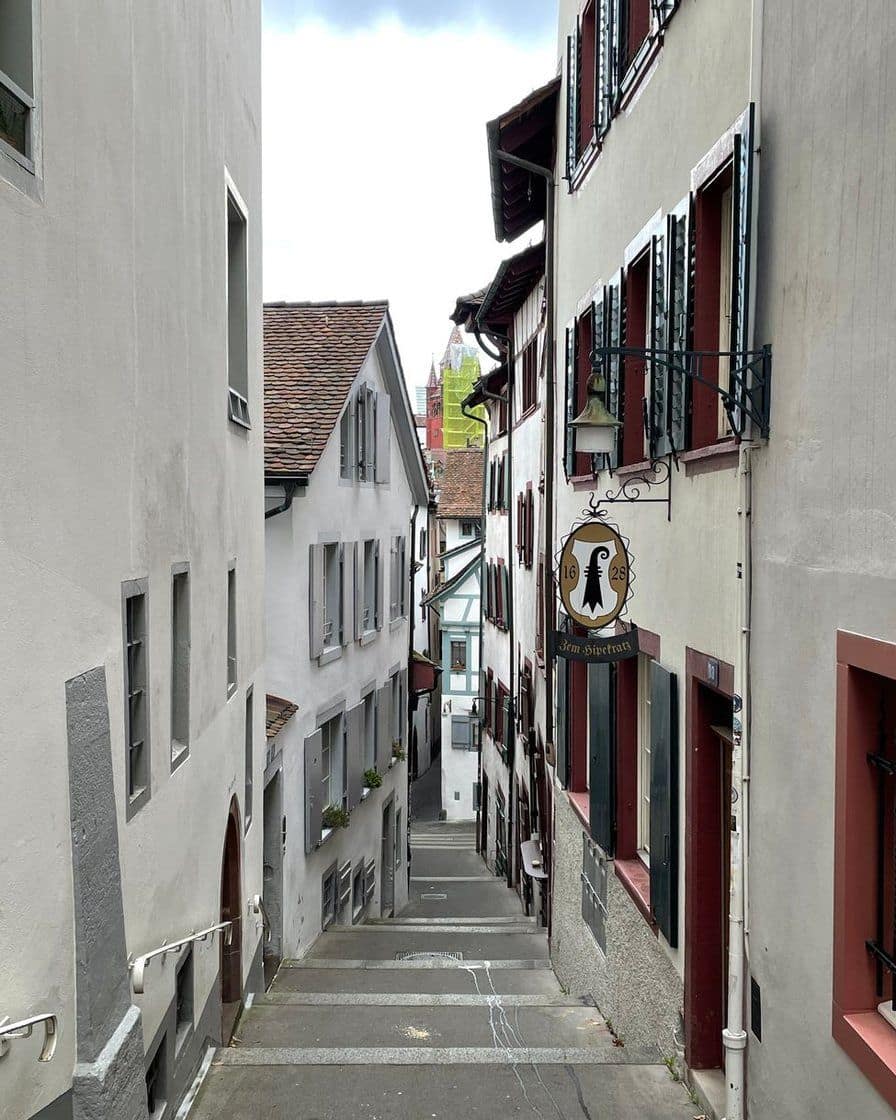 Eine steile Treppengasse in der Basler Altstadt, weisse Häuser mit roten Fensterrahmen und ein Wirtshausschild mit dem Baselstab, im Hintergrund ein roter Kirchturm
