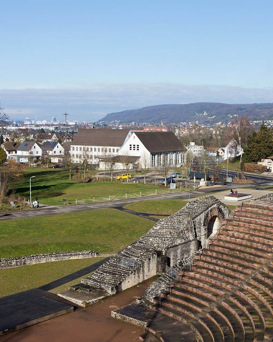 Das römische Theater von Augusta Raurica bei Basel, halbrunde Steinstufen und Mauerreste auf einer grünen Wiese unter Bäumen