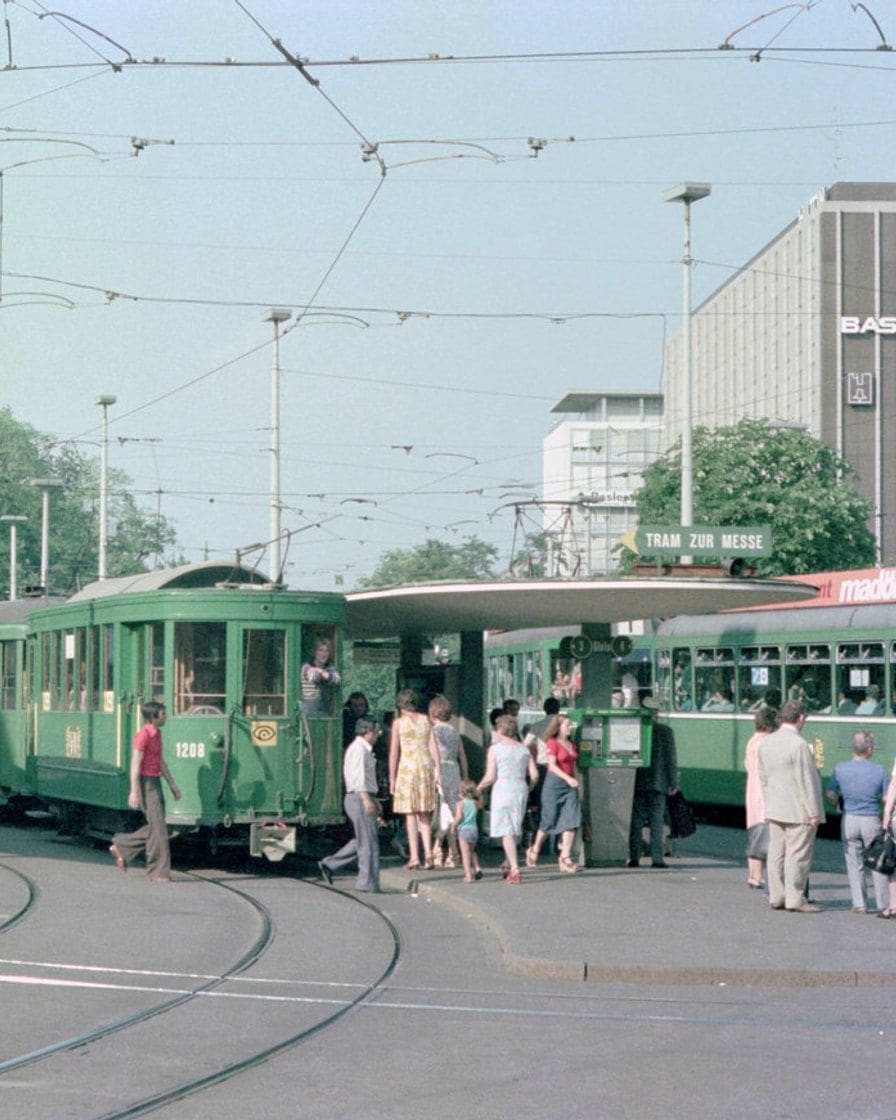 Die Tramhaltestelle vor dem Bahnhof SBB in Basel mit wartenden Fahrgästen, Anzeigetafel und zwei Trams nebeneinander