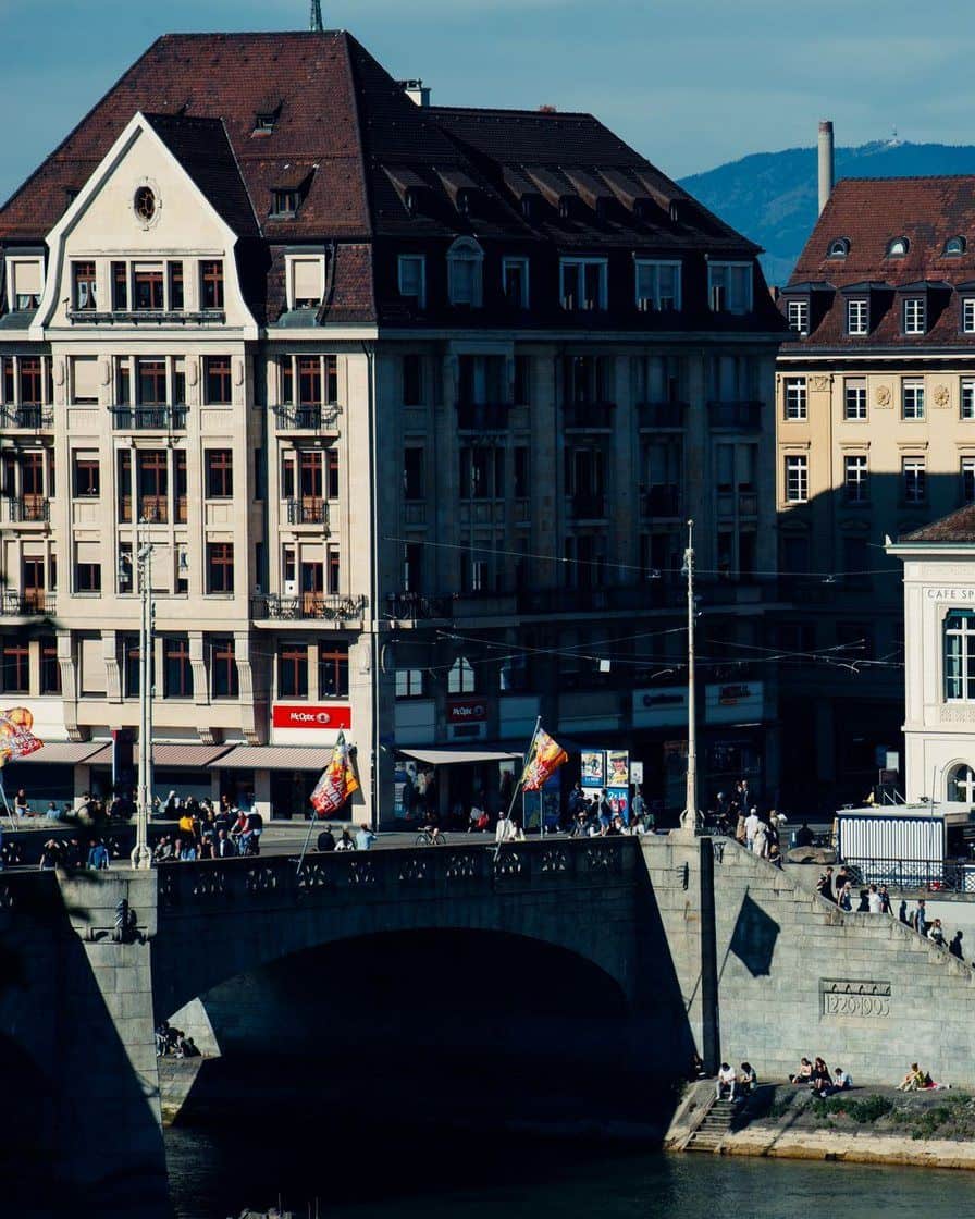 Der Kleinbasler Brückenkopf der Mittleren Brücke an einem sonnigen Tag, Menschen sitzen unten am Rheinufer, auf der Brücke wehen Fahnen