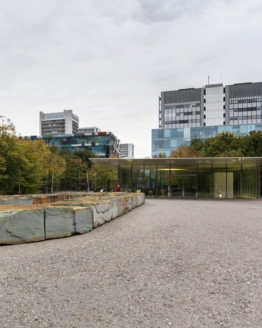 Le Novartis Campus à Bâle-St. Johann avec ses façades modernes de verre et de métal le long d'une promenade arborée, des passants sur l'allée