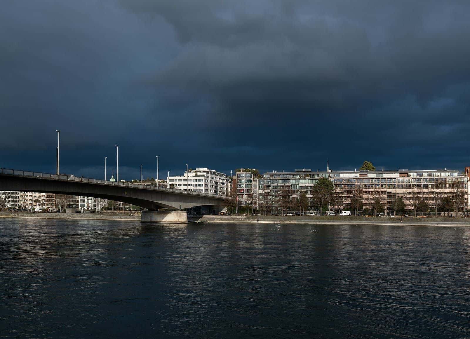 Der Untere Rheinweg in Kleinbasel unter einem dunklen Gewitterhimmel, im Hintergrund die Johanniterbrücke über den Rhein